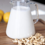 Glass pitcher filled with creamy homemade cashew milk on a wooden board with raw cashews nearby — easy no-strain dairy-free milk recipe.