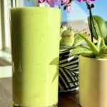 Tall glass of creamy green smoothie on a sunny windowsill with a lemon, green apple, and avocado beside it