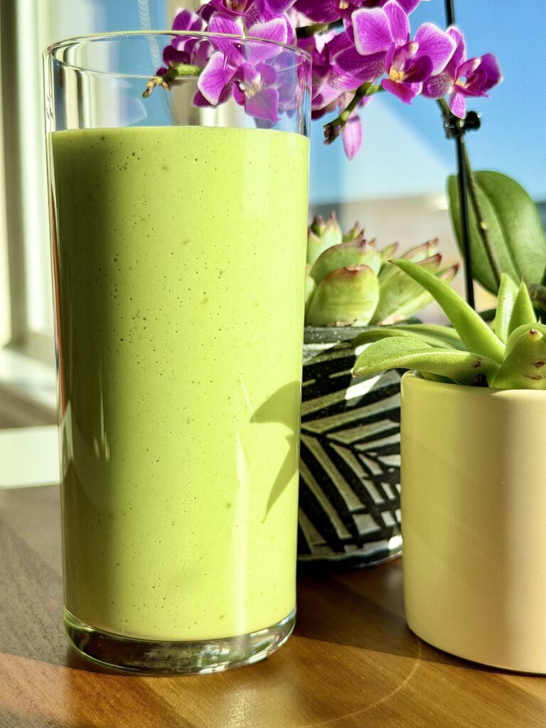 Tall glass of creamy green smoothie on a sunny windowsill with a lemon, green apple, and avocado beside it