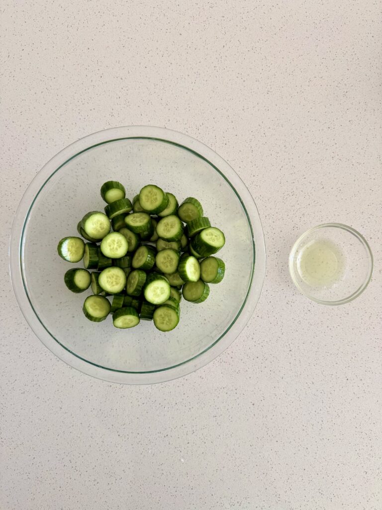 Salted cucumber slices in a glass bowl next to a small bowl of drained cucumber liquid