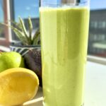 Tall glass of creamy green smoothie on a sunny windowsill with a lemon, green apple, and avocado beside it