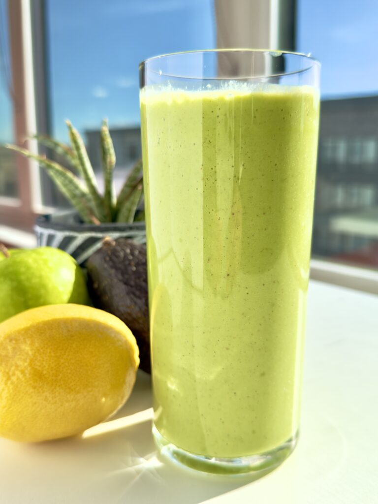 Tall glass of creamy green smoothie on a sunny windowsill with a lemon, green apple, and avocado beside it