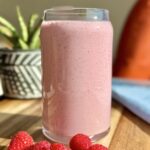 Creamy pink raspberry protein smoothie in a clear glass with fresh raspberries on a wooden board