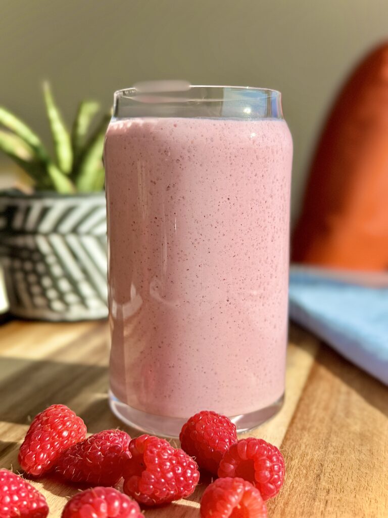Creamy pink raspberry protein smoothie in a clear glass with fresh raspberries on a wooden board