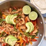 Skillet chicken fajitas with bell peppers, onions, avocado, and lime in a stainless-steel pan