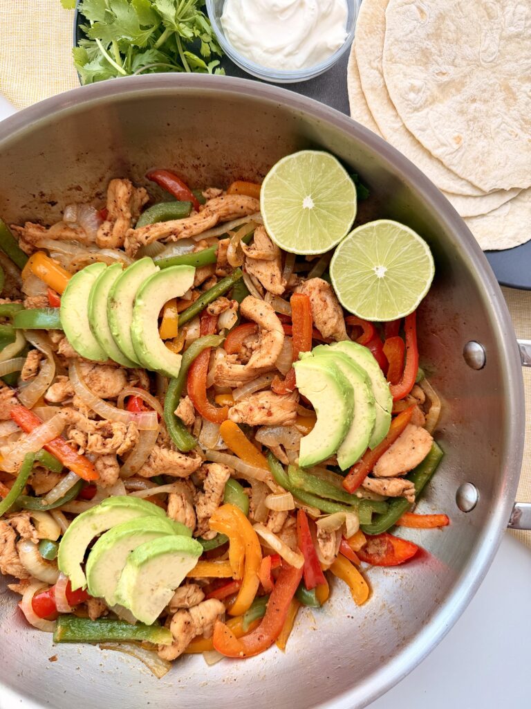 Skillet chicken fajitas with bell peppers, onions, avocado, and lime in a stainless-steel pan