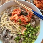Korean bone broth noodle soup with tender beef, scallions, and kimchi in a white bowl