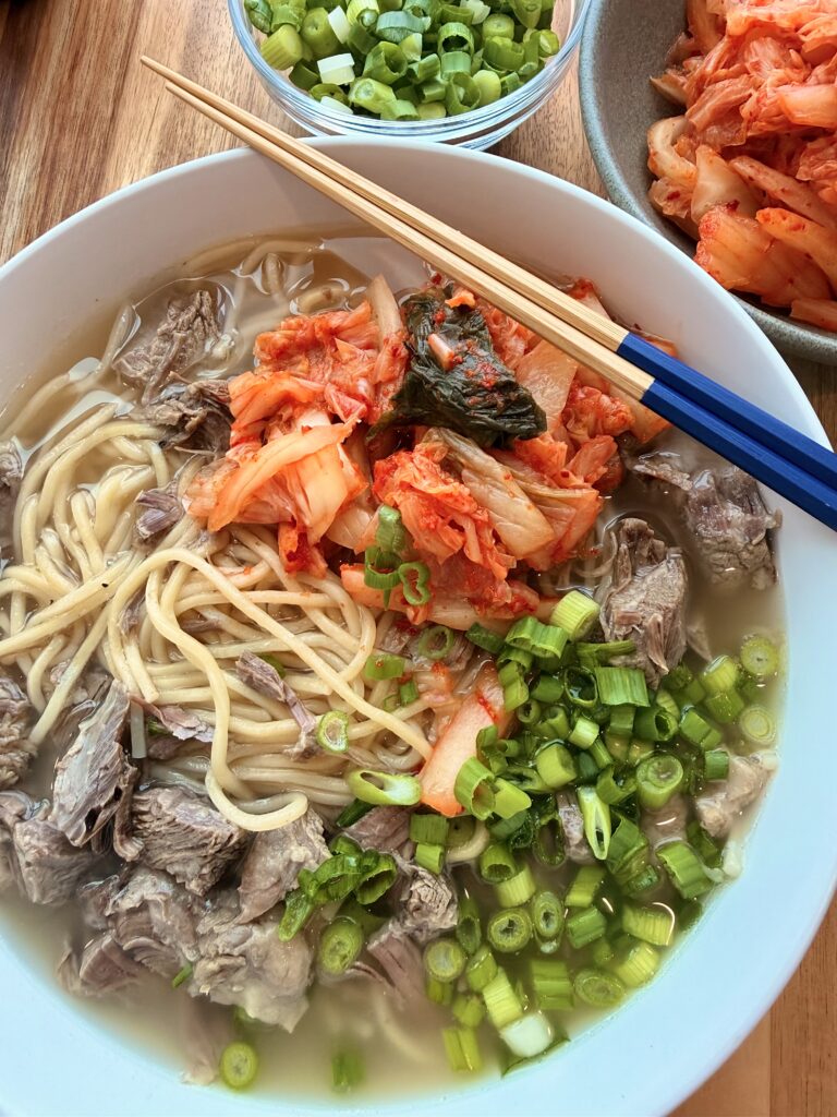 Korean bone broth noodle soup with tender beef, scallions, and kimchi in a white bowl