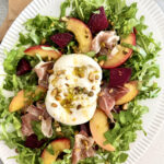Summer Peach Burrata Salad with prosciutto, pistachios, roasted beets, arugula, basil, and white balsamic vinaigrette on a serving platter.