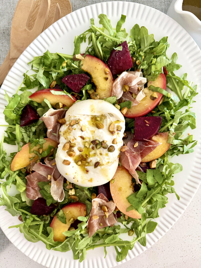 Summer Peach Burrata Salad with prosciutto, pistachios, roasted beets, arugula, basil, and white balsamic vinaigrette on a serving platter.