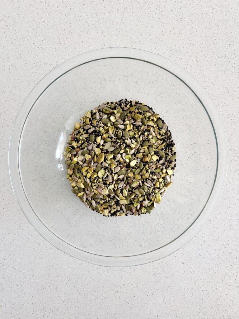 Black and white sesame seeds, chopped pistachios, pumpkin seeds, sunflower seeds, and hemp hearts combined in a glass bowl.