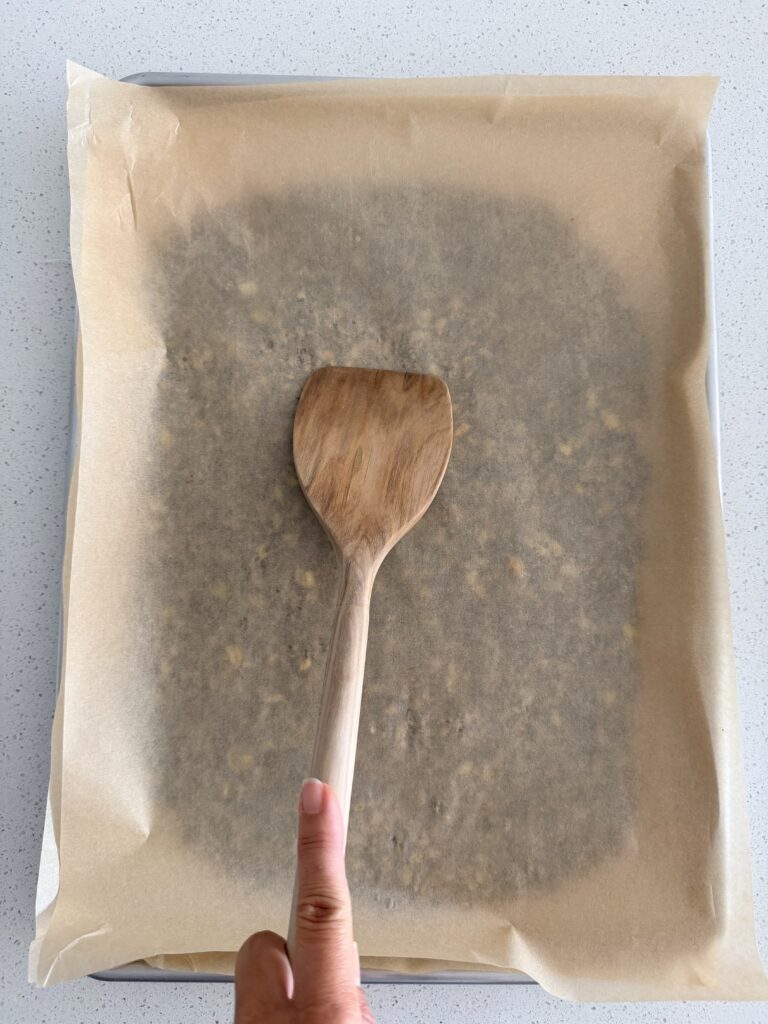 Wooden spatula pressing the seed mixture flat under a second sheet of parchment on a baking sheet.