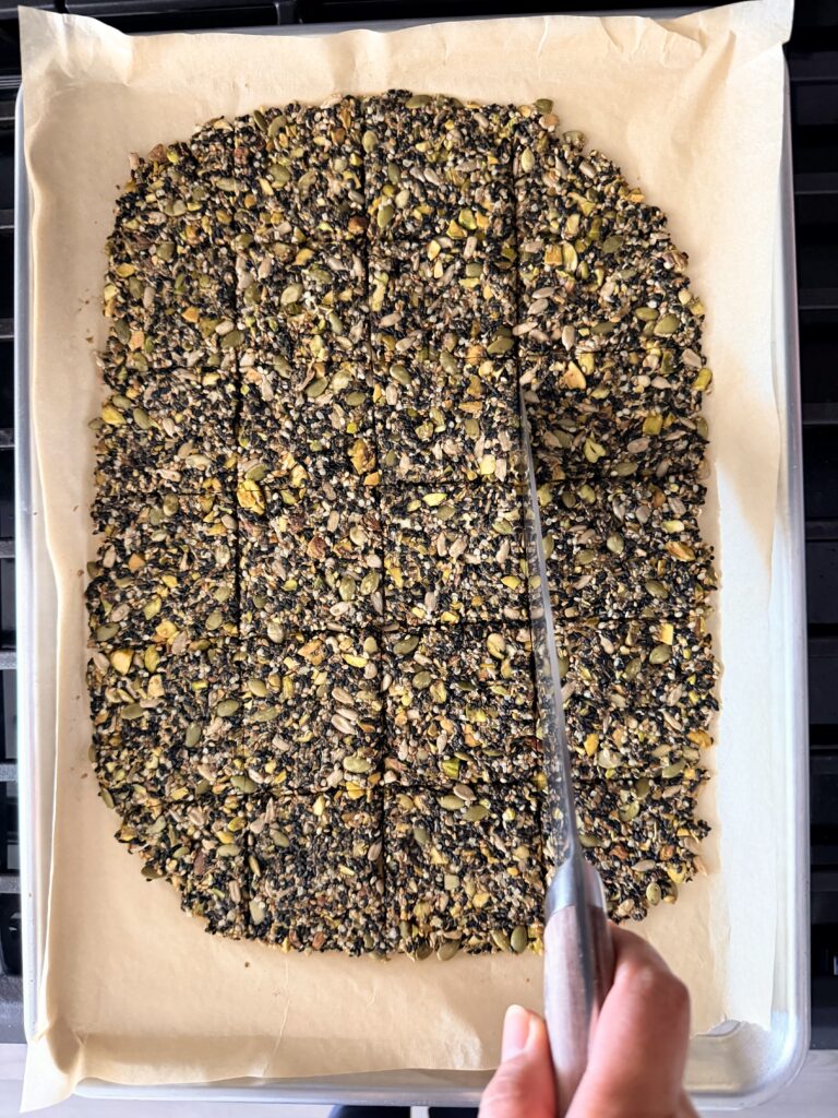 Knife scoring partially baked black sesame seed crackers into squares on a parchment-lined baking sheet.