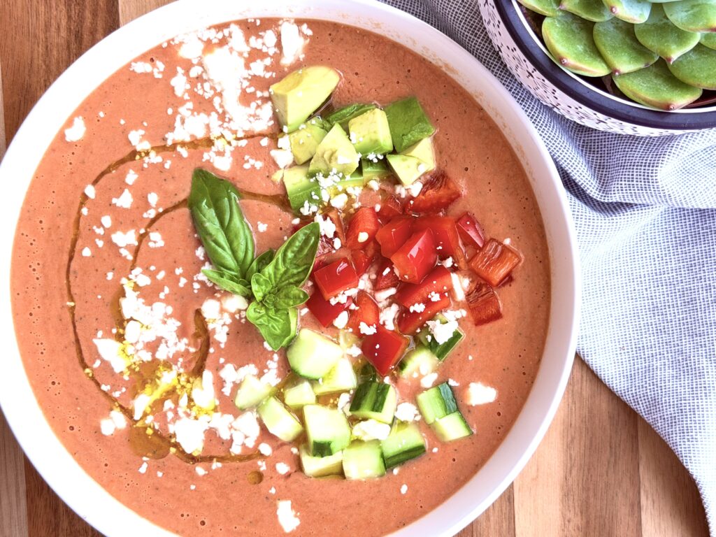 Overhead shot of creamy high-protein gazpacho in a white bowl, topped with avocado, red bell pepper, cucumber, feta, basil, and a swirl of olive oil