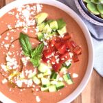 Overhead shot of creamy high-protein gazpacho in a white bowl, topped with avocado, red bell pepper, cucumber, feta, basil, and a swirl of olive oil