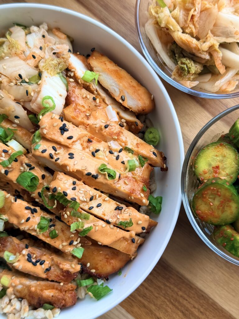 Close-up of sliced gochujang chicken drizzled with creamy gochujang yogurt sauce over farro with kimchi and cucumber salad.