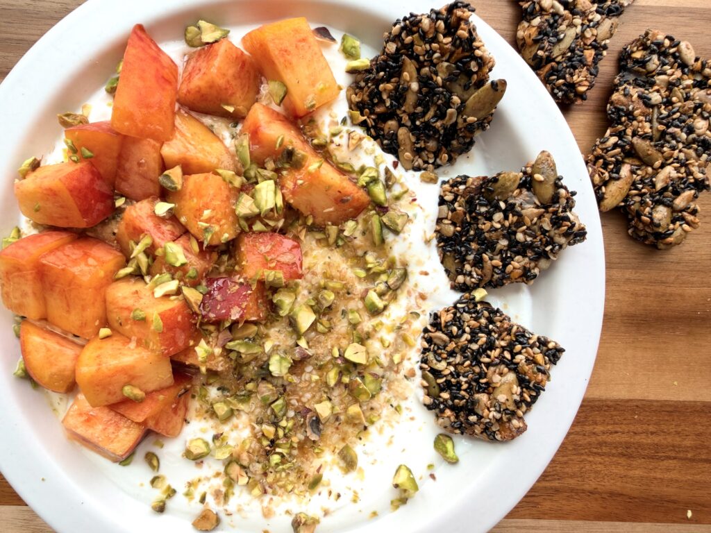 Peach Greek yogurt bowl with hemp hearts, ground flaxseed, fresh peaches, roasted pistachios, honey, and Healthy Black Sesame Seed Crackers, with extra homemade seed crackers alongside.