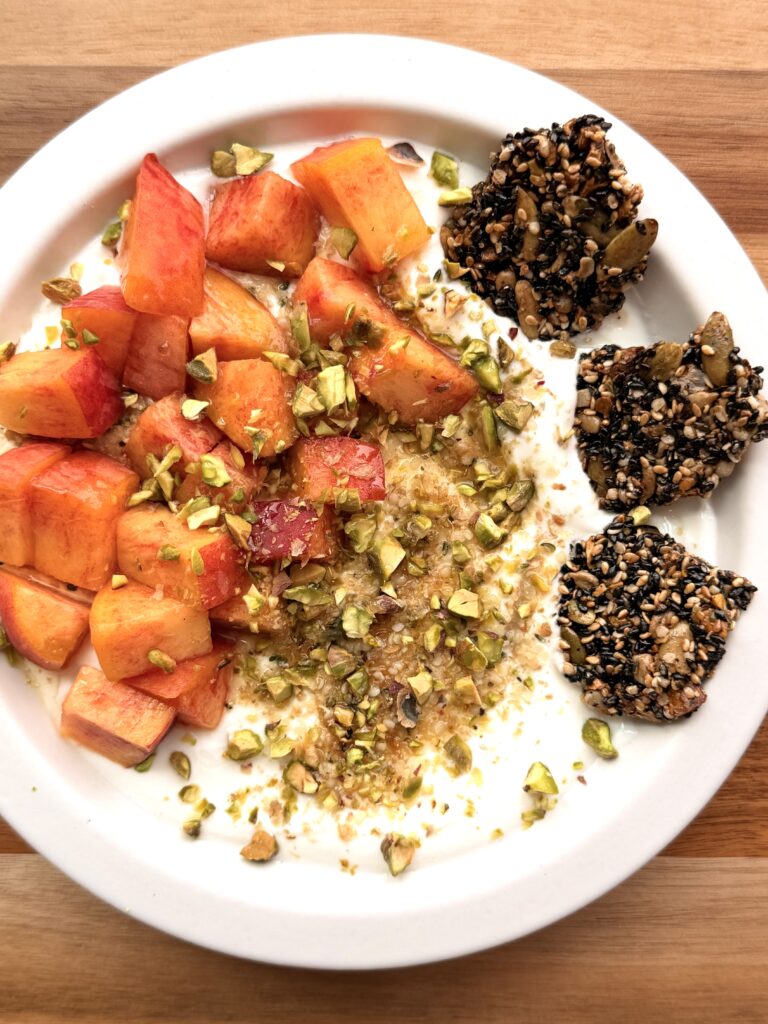 Peach Greek yogurt bowl with hemp hearts, ground flaxseed, fresh peaches, roasted pistachios, honey, and Healthy Black Sesame Seed Crackers.