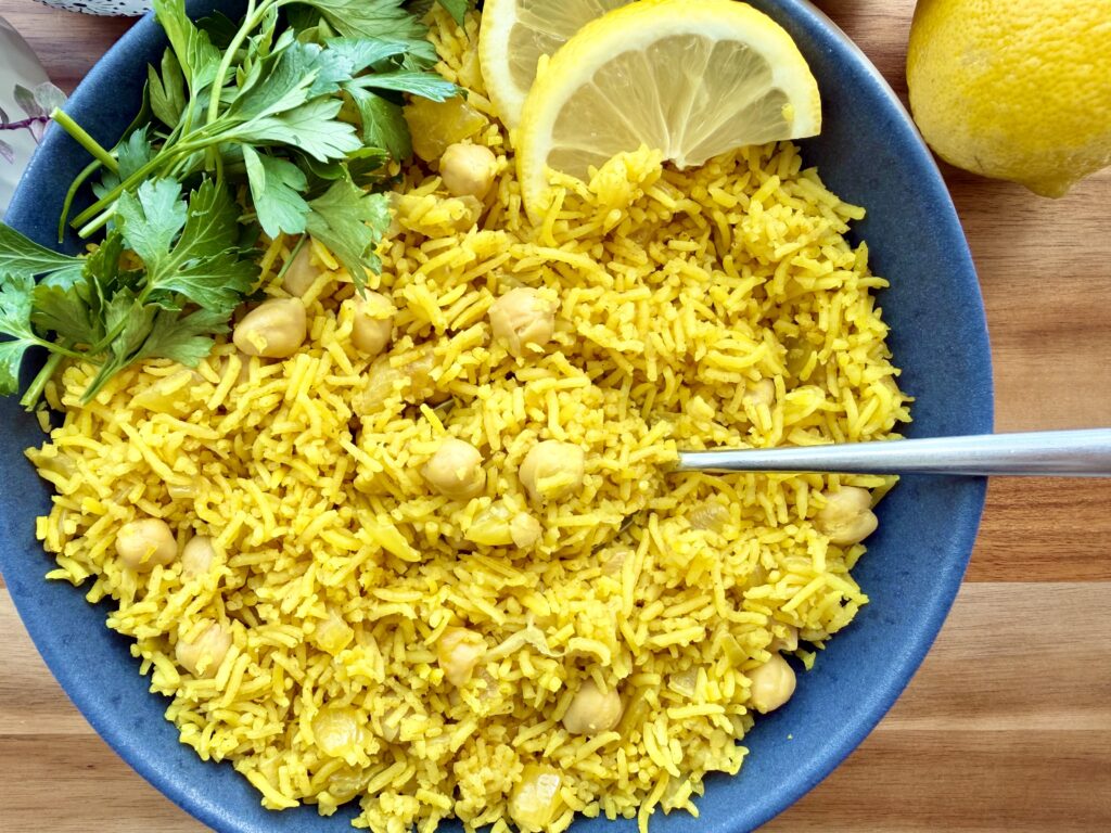 Overhead shot of golden turmeric rice with chickpeas in a blue bowl, garnished with fresh parsley and lemon wedges