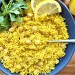 Overhead shot of golden turmeric rice with chickpeas in a blue bowl, garnished with fresh parsley and lemon wedges