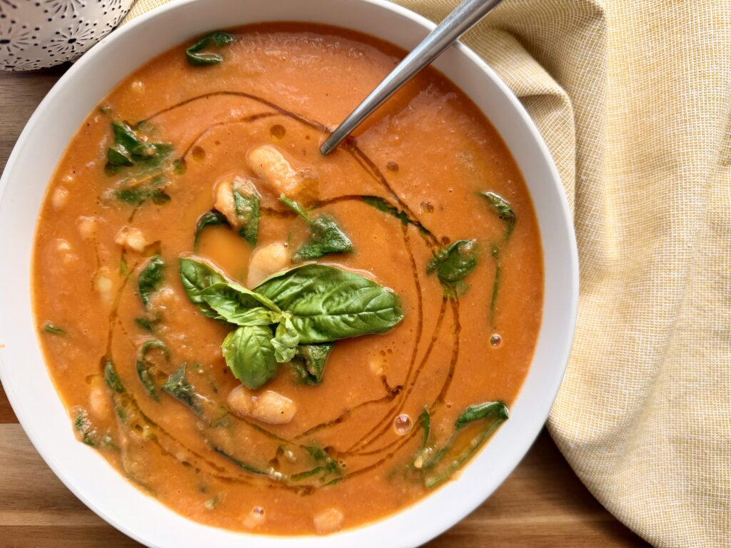 Creamy roasted tomato and white bean soup with wilted spinach and fresh basil in a white bowl on a wooden table