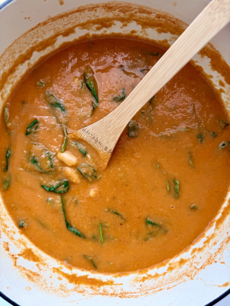 Baby spinach and whole cannellini beans stirred into creamy roasted tomato soup with a wooden spoon