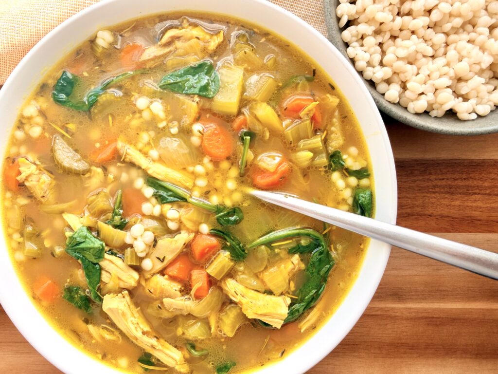 Bowl of ginger turmeric chicken soup with shredded chicken, carrots, spinach, and pearl couscous on a wooden board, with extra couscous on the side