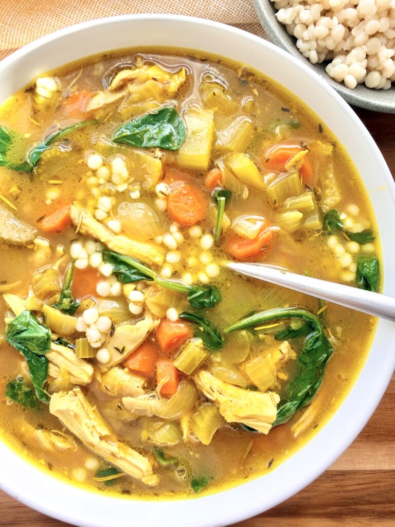 Close-up of golden ginger turmeric chicken soup with pearl couscous, shredded chicken, carrots, celery, and baby spinach