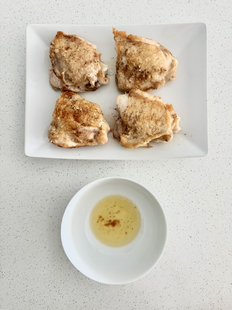 Seared chicken thighs on a white platter next to a bowl of rendered chicken fat spooned from the pot