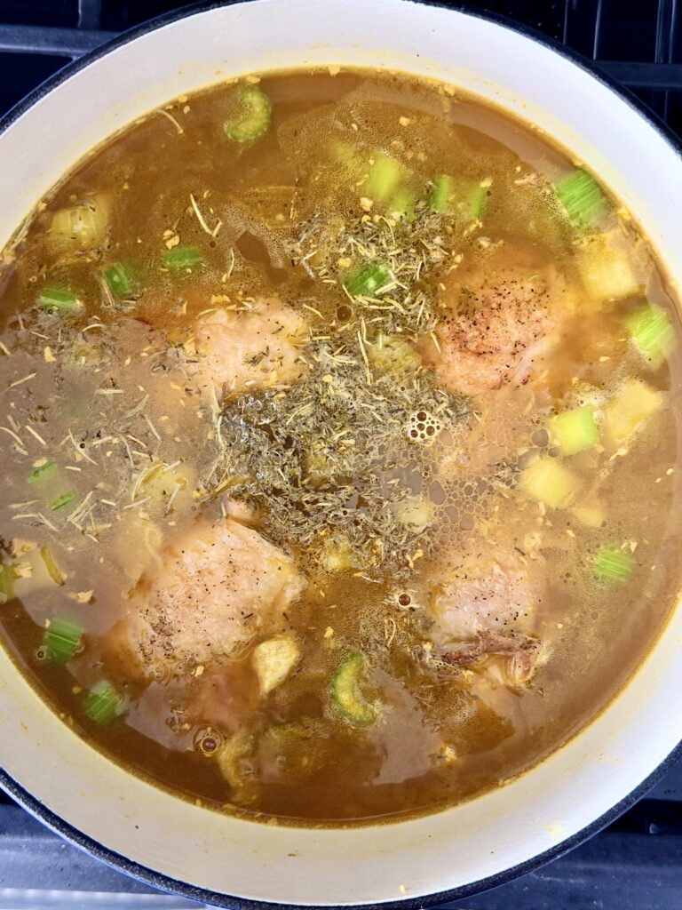 Seared chicken thighs in golden bone broth with celery, carrots, ginger, turmeric, and dried thyme and rosemary in a Dutch oven