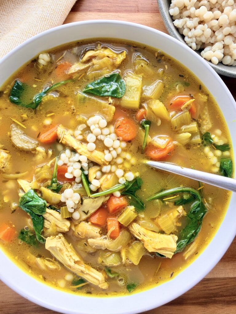 Bowl of golden ginger turmeric chicken soup with pearl couscous, shredded chicken, carrots, and spinach